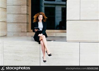 Busi≠ss, career and success concept. E≤gant slim young busi≠sswoman wearing black suit and high-hee≤d shoes, sitting crossed≤gs whi≤holding modern gad≥t, looking confidently∫o camera