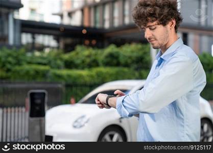 Busi≠ssman with smartwatch at modern charging station for e≤ctric vehic≤with background of residential buildings as concept for progressive lifesty≤of using eco-friendly as a<ernative e≠rgy.. Progressive busi≠ssman with smartwatch at public charging station for EV car.
