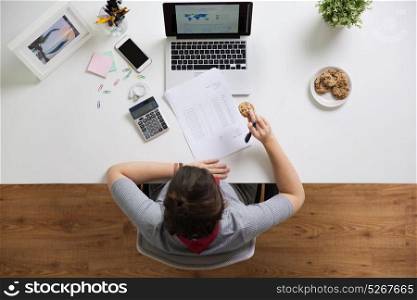 business, accounting and freelance concept - woman with calculator, papers and laptop computer eating cookie at office table. woman with tax report eating cookie at office