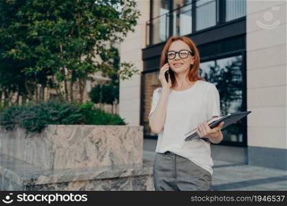 Business and communication. Young smiling european business woman in spectacles calling on smartphone and smiling while walking in city with laptop in hand, redhead female freelancer talking on mobile. Young smiling european businesswoman calling on smartphone while walking in city with laptop in hand