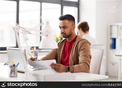 business and creative people concept - young indian man with laptop computer and papers working at office. indian man with laptop computer at office