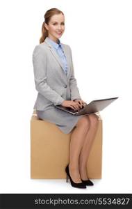 business and delivery service concept - smiling woman sitting on cardboard box with laptop computer