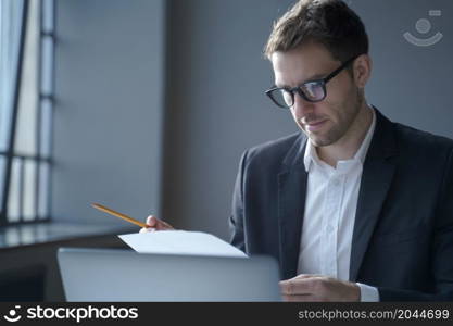 Business and job concept. Serious male professional analyzing documents at workplace, working on laptop computer in office, focused german businessman wearing glasses and black suit. Serious male professional analyzing documents at workplace. Business and job concept