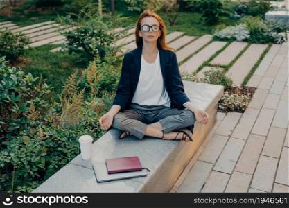 Business and meditation concept. Redhead young European woman sits in lotus position wears formal clothes drinks takeaway coffee uses notepad digital device does yoga asana poses outside in park. Redhead young European woman sits in lotus position wears formal clothes drinks takeaway coffee