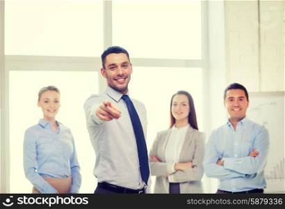 business and office concept - smiling handsome businessman with team in office pointing finger at you. businessman in office pointing finger at you
