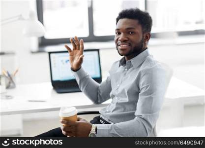 business and people concept - african american businessman with coffee sitting at office table and waving hand. african american businessman with coffee at office. african american businessman with coffee at office