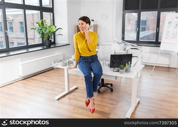 business and people concept - happy businesswoman sitting on desk and calling on smartphone at office. happy woman and calling on smartphone at office