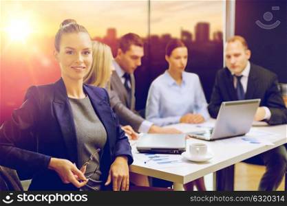business and people concept - smiling businesswoman with eyeglasses and team at office over city background. businesswoman and business team at office