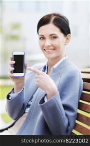 business and people concept - young smiling businesswoman showing smartphone blank screen and sitting on city bench. young smiling businesswoman showing smartphone