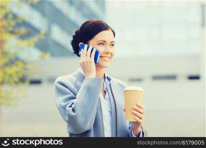 business and people concept - young smiling woman calling on smartphone and drinking coffee over office building in city. smiling woman with coffee calling on smartphone
