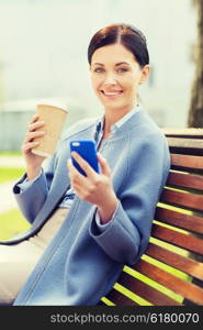 business and people concept - young smiling woman with smartphone sitting on bench and drinking coffee in city. smiling woman with coffee and smartphone