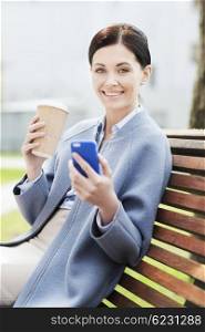 business and people concept - young smiling woman with smartphone sitting on bench and drinking coffee in city. smiling woman with coffee and smartphone