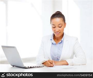 business and technology concept - african businesswoman with laptop writing something in notebook