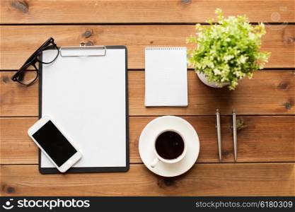 business and technology concept - close up of smartphone, clipboard, coffee cup, eyeglasses and notepad on wooden table