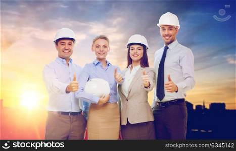 business, architecture and building concept - happy team of architects in white hard hats showing thumbs up over city background. business team in white hard hats showing thumbs up