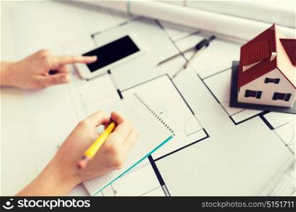 business, architecture, building, construction and people concept - close up of architect hand with living house blueprint and smarphone writing to notebook. close up of hand on blueprint writing to notebook