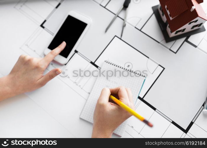 business, architecture, building, construction and people concept - close up of architect hand with living house blueprint and smarphone writing to notebook