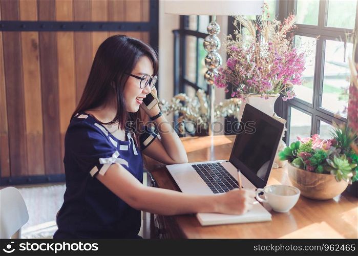 Business Asian woman hold pick up Smartphone make a note and working with laptop computer on in coffee shop like the background.