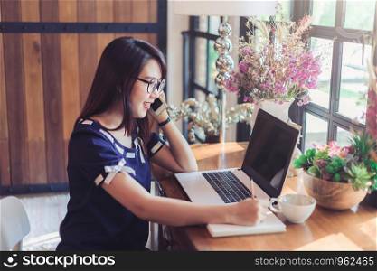 Business Asian woman hold pick up Smartphone make a note and working with laptop computer on in coffee shop like the background.
