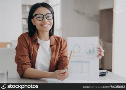 Business assistant points to the statistical data graph with pencil. Happy young spanish manager in glasses is working remote at home workspace. Attractive girl is doing paperwork and smiling.. Business assistant points to the statistical data graph with pencil. Paperwork at home.