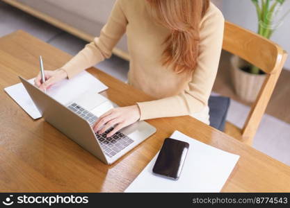 Business at home concept, Young Asian woman is reading data on laptop and taking notes on notebook.