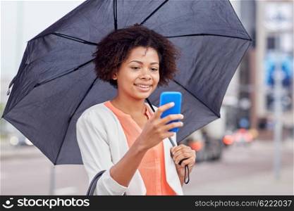 business, bad weather and people and concept - happy african american businesswoman with umbrella texting on smartphone at city street