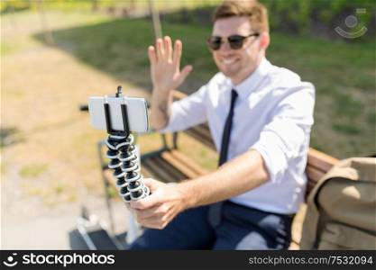 business, blogging and technology concept - young businessman recording video blog by smartphone on tripod and waving hand in city. businessman recording video blog by smartphone