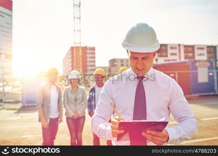 business, building, teamwork and people concept - group of smiling builders and architect with clipboard in hardhats at construction site