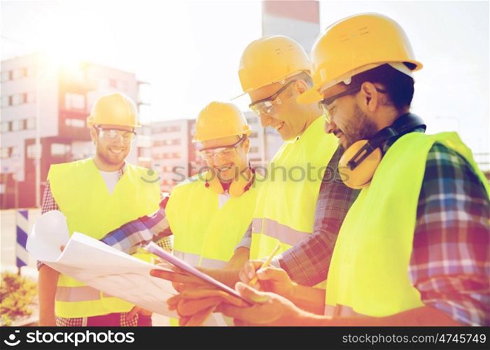 business, building, teamwork and people concept - group of smiling builders in hardhats with clipboard and blueprint outdoors
