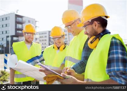 business, building, teamwork and people concept - group of smiling builders in hardhats with clipboard and blueprint outdoors