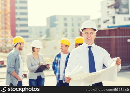 business, building, teamwork and people concept - group of smiling builders in hardhats with clipboard and blueprint outdoors