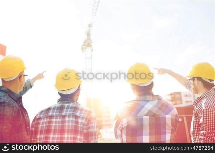 business, building, teamwork, development and people concept - group of builders in hardhats at construction site pointing finger to crane from back