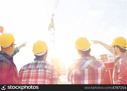 business, building, teamwork, development and people concept - group of builders in hardhats at construction site pointing finger to crane from back