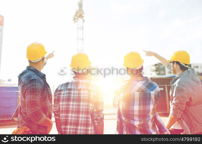 business, building, teamwork, development and people concept - group of builders in hardhats at construction site pointing finger to crane from back