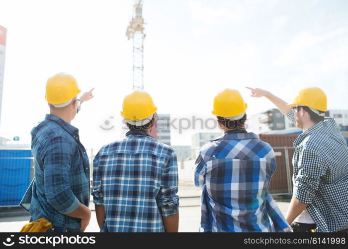 business, building, teamwork, development and people concept - group of builders in hardhats at construction site pointing finger to crane from back