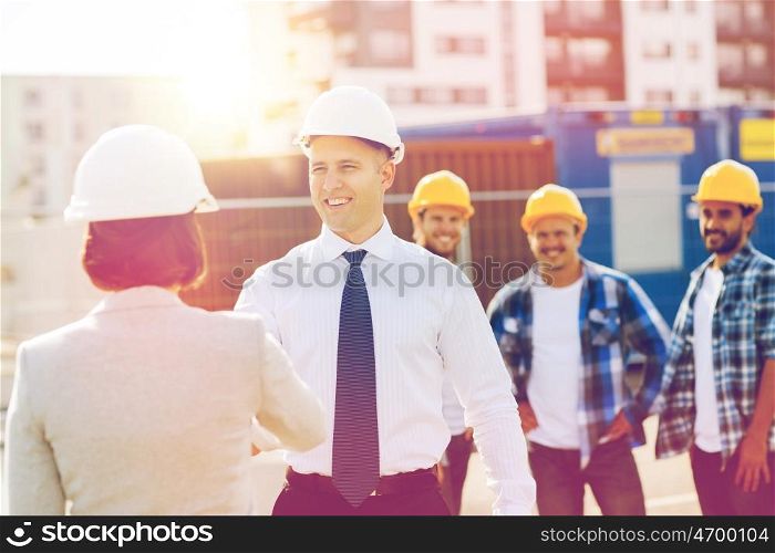 business, building, teamwork, gesture and people concept - group of smiling builders in hardhats with clipboard greeting each other outdoors