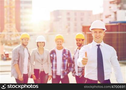 business, building, teamwork, gesture and people concept - group of smiling builders in hardhats showing thumbs up outdoors