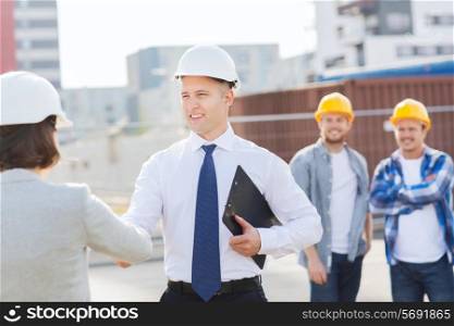 business, building, teamwork, gesture and people concept - group of smiling builders in hardhats with clipboard greeting each other outdoors