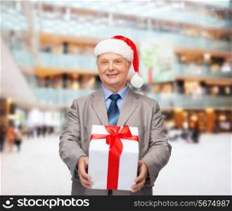 business, christmas, presents and people concept - smiling senior man in suit and santa helper hat with gift over shopping center background