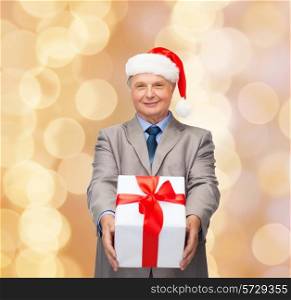 business, christmas, presents and people concept - smiling senior man in suit and santa helper hat with gift over beige lights background