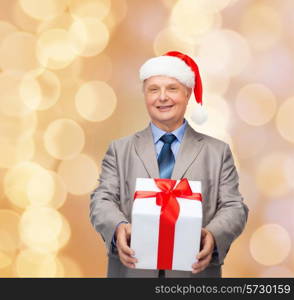 business, christmas, presents and people concept - smiling senior man in suit and santa helper hat with gift over beige lights background