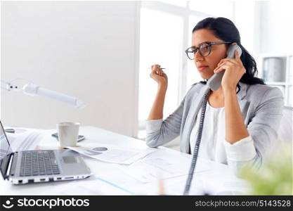 business, communication and people concept - businesswoman with laptop computer and papers calling on desk phone at office. businesswoman calling on desk phone at office