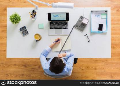 business, communication and people concept - businesswoman with laptop computer calling on phone and writing to notebook at office table. businesswoman calling on phone at office table