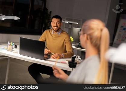 business, communication and people concept - creative man or graphic designer with laptop computer working by pen tablet and talking to female colleague at night office. designer with laptop talking to coworker at office. designer with laptop talking to coworker at office