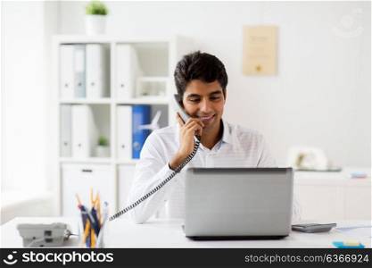 business, communication and people concept - happy businessman with laptop computer calling on desk phone at office. happy businessman calling on desk phone at office