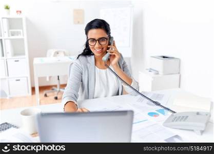 business, communication and people concept - happy smiling businesswoman with laptop computer and papers calling on desk phone at office. businesswoman calling on desk phone at office