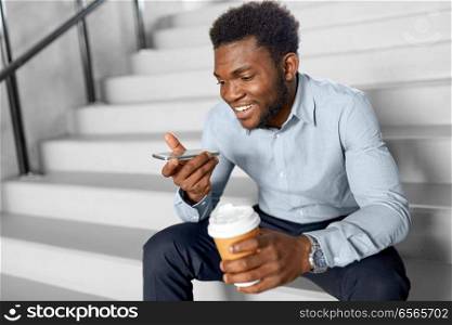 business, communication and technology concept - african american businessman calling or using voice recorder on smartphone at coffee break on office stairs. businessman records voice by smartphone at office