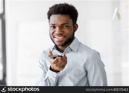 business, communication and technology concept - african american businessmanr calling or using voice recorder on smartphone at office. businessman records voice by smartphone at office. businessman records voice by smartphone at office