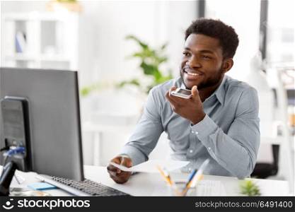 business, communication and technology concept - african americanbusinessman with papers and computer calling or using voice recorder on smartphone at office. businessman records voice by smartphone at office. businessman records voice by smartphone at office