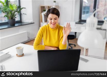business, communication and technology concept - businesswoman having video call on laptop computer and waving hand at office. businesswoman having video call at office